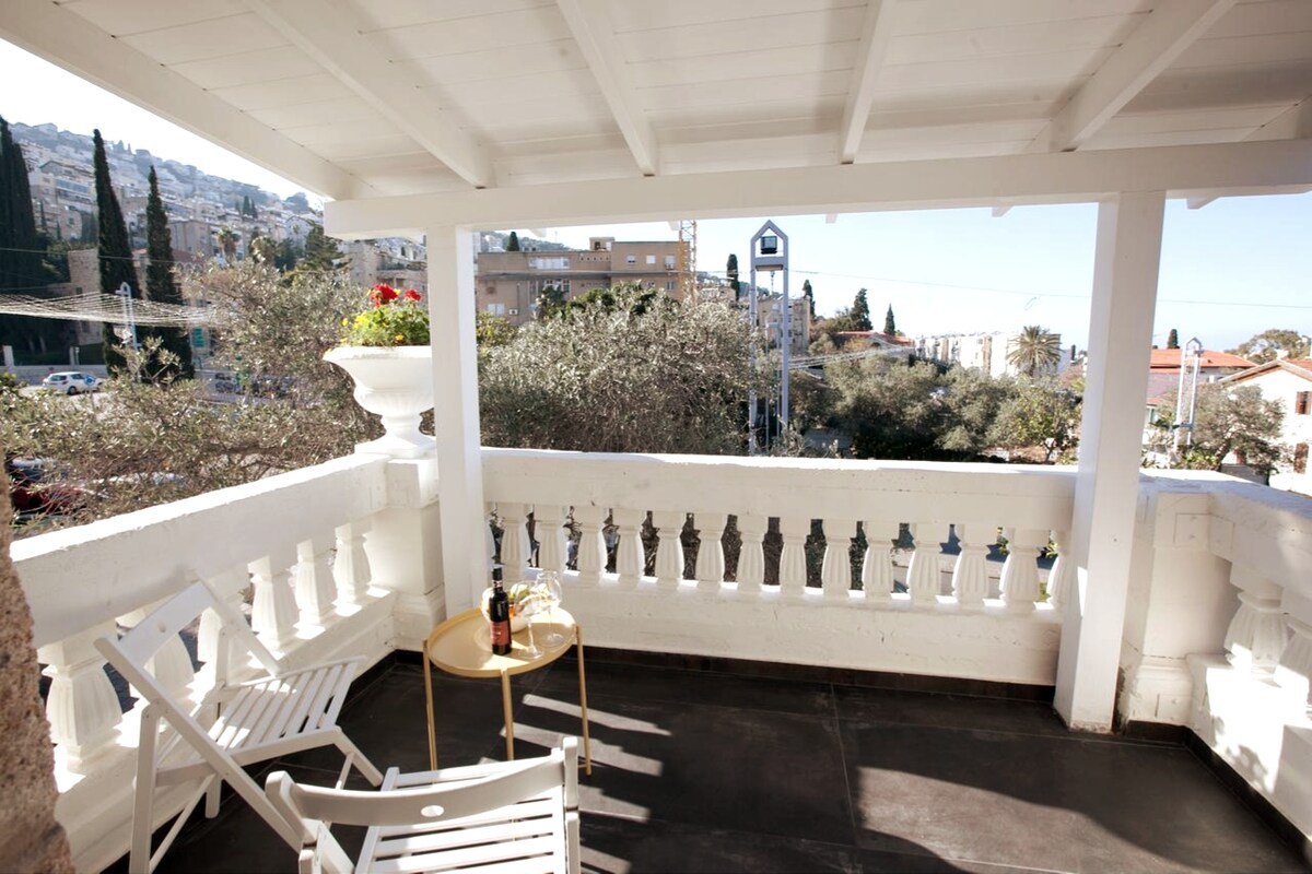 A covered balcony features two white lounge chairs positioned next to a small round table. Potted flowers add a touch of color. The view encompasses trees and buildings in the bustling neighborhood, allowing for a glimpse of the lively surroundings.