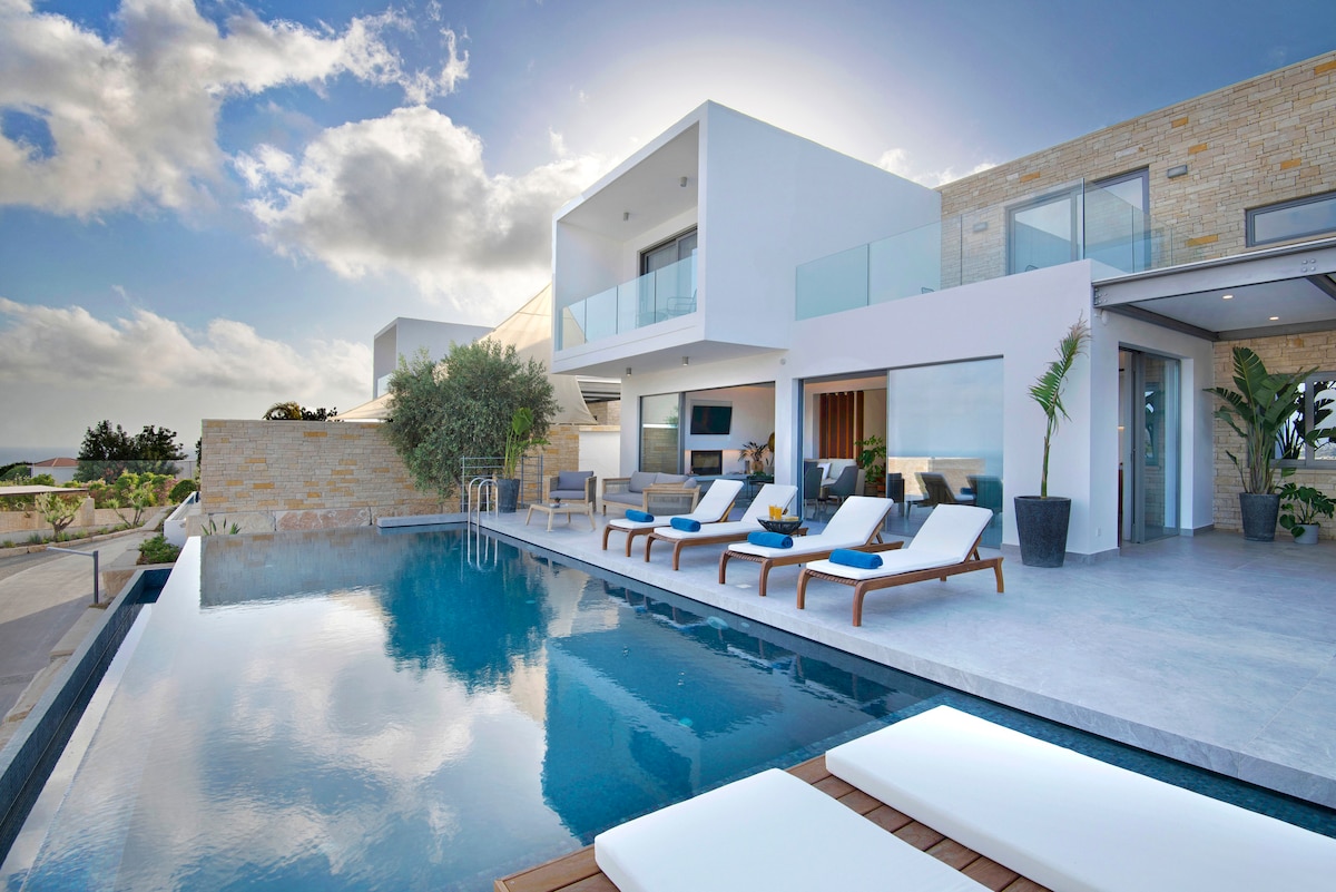 An outdoor area features an infinity pool reflecting the sky and surrounding landscape. Comfortable sun loungers are arranged along the poolside, with a modern seating area visible in the background. Large glass doors provide access to the interior, enhancing the open-air ambiance.