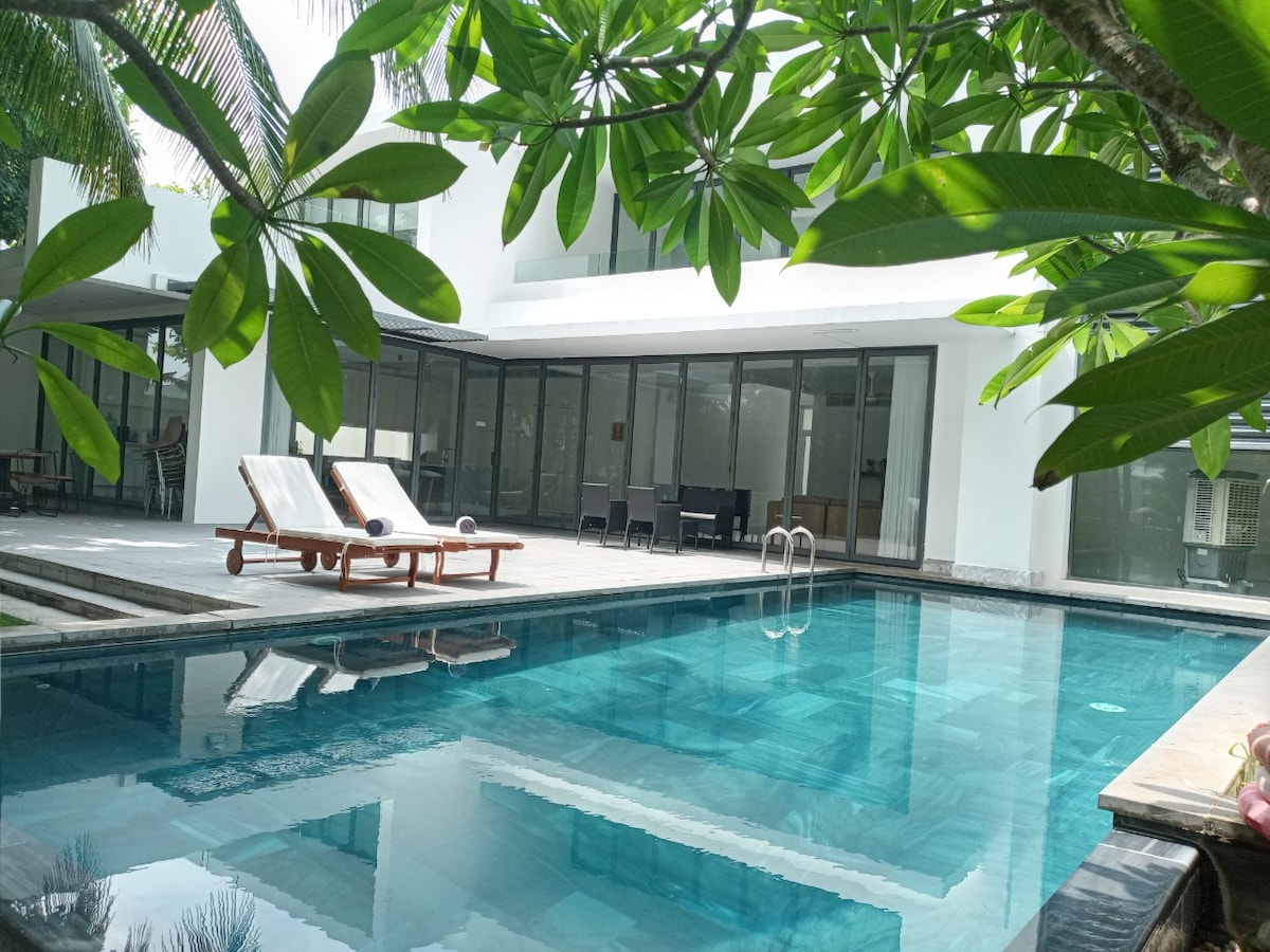 A crystal-clear private pool is framed by lush green foliage, reflecting the vibrant surroundings. Sun loungers are positioned on the wooden deck beside the pool, while large glass doors lead into the open-plan living area of the villa, enhancing the tranquil ambiance.