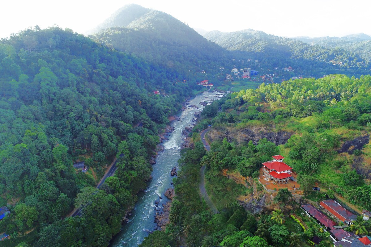 A sweeping aerial view captures the river winding through lush greenery, surrounded by hills. The vibrant landscape features a cluster of houses, with one prominently situated near the riverbank, offering scenic vistas of the valley and waterway below.