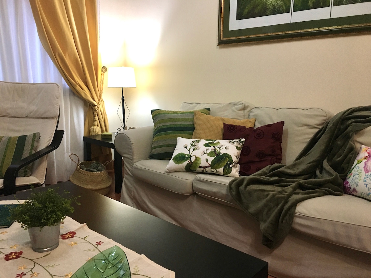 A comfortable living area is presented with a light-colored sofa adorned with various decorative pillows. A dark coffee table sits in front, complemented by a small plant. Soft lighting is provided by a lamp, while golden curtains frame a nearby window.