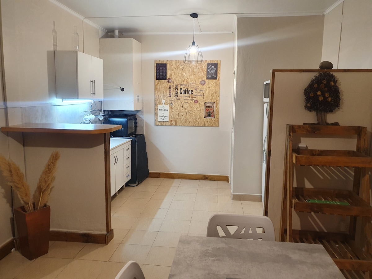 A well-equipped kitchen space is featured, showcasing a minimalist design with white cabinetry and a rustic bulletin board. A small table with chairs is positioned in the foreground, while decorative plants add a natural touch to the ambiance.