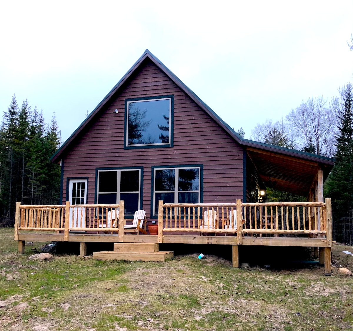 The rustic cabin features a wooden exterior with large windows, allowing natural light to fill the interior. A spacious wrap-around deck is surrounded by trees, equipped with simple outdoor chairs, inviting guests to relax in the serene setting.