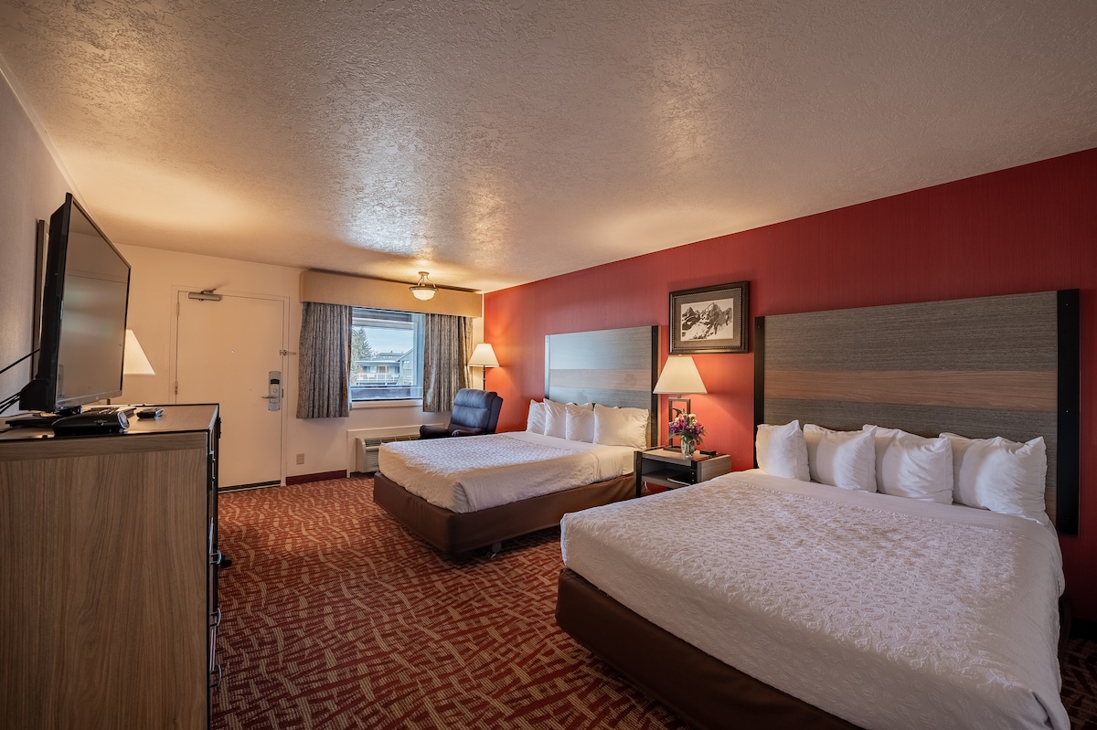 The room is furnished with two spacious beds, a large television, and a comfortable armchair. Natural light enters through a window covered with patterned curtains. The decor includes a framed piece of artwork, and the flooring features a warm carpet design.