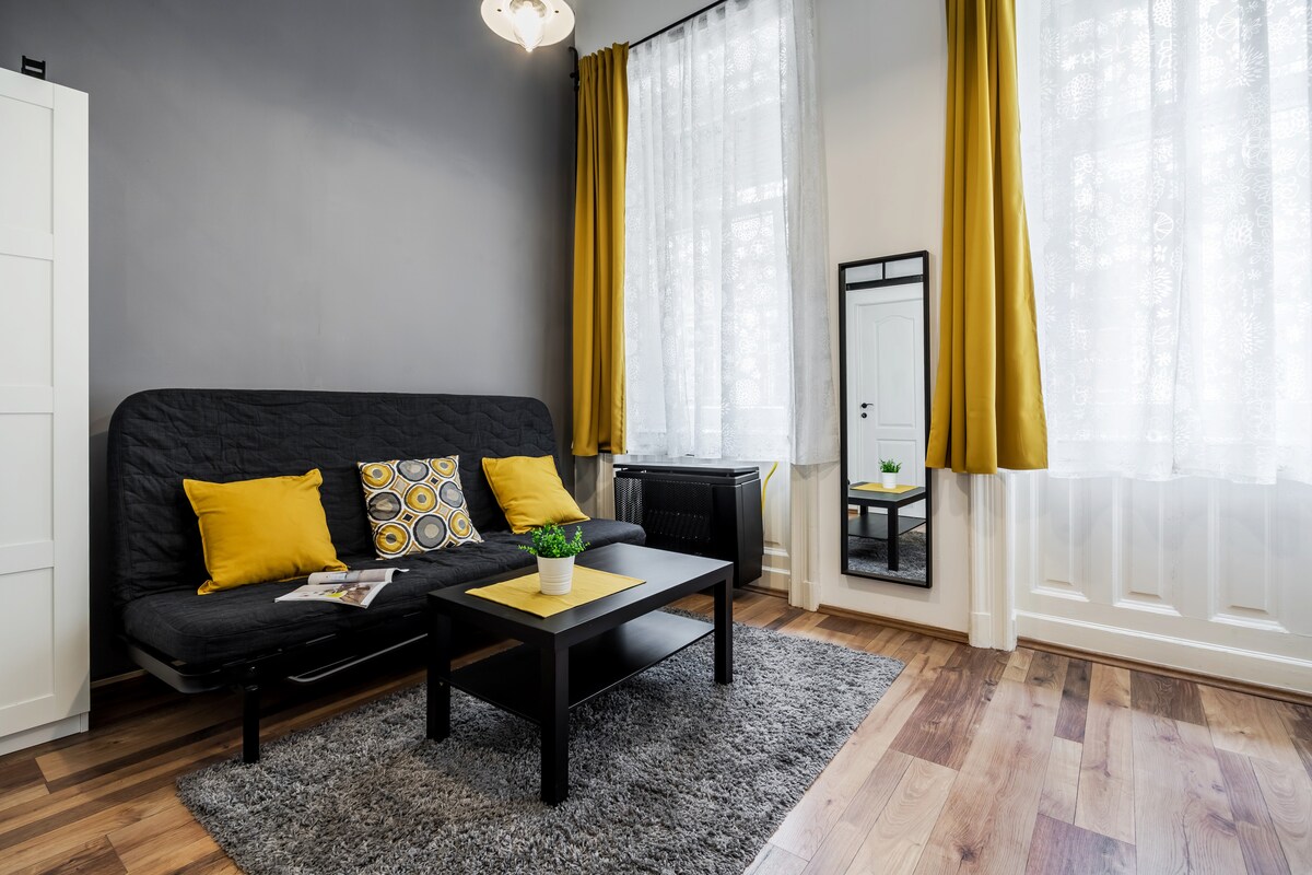 A stylish living area features a black sofa with bright yellow cushions, complemented by a coffee table on a textured rug. Natural light fills the space through large windows adorned with sheer curtains. A mirror and decorative plant add subtle elegance to the setting.