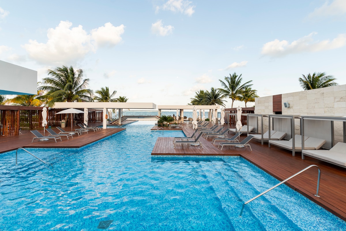 A spacious pool area is surrounded by sun loungers and private cabanas, providing opportunities for relaxation. Lush palm trees line the perimeter, and the clear blue water sparkles under a bright sky, adding to the inviting atmosphere of the space.