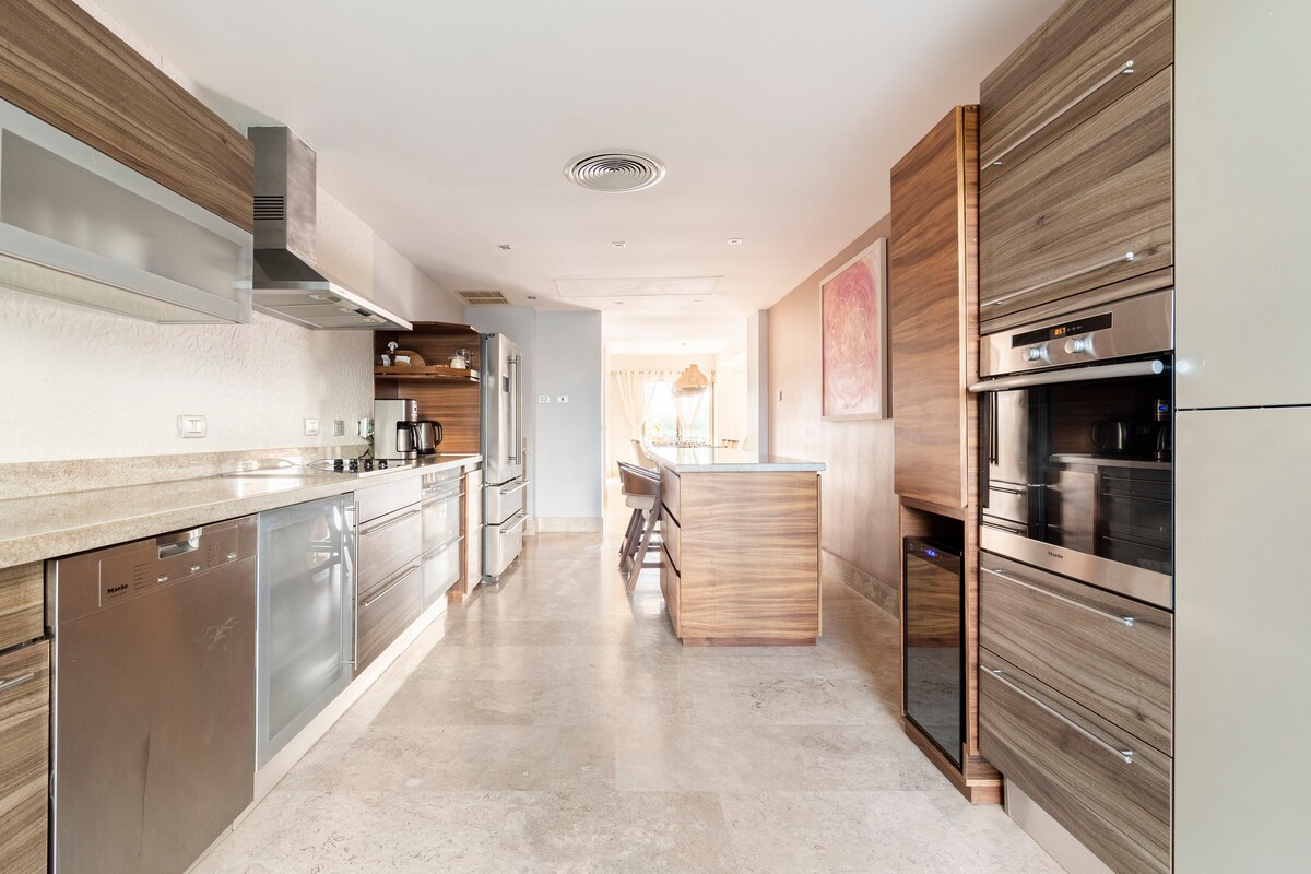An open-concept kitchen showcases modern appliances and sleek cabinetry in warm wood tones. A spacious island provides additional preparation space, while natural light flows in from nearby windows. The kitchen area is seamlessly connected to the dining space, enhancing functionality.