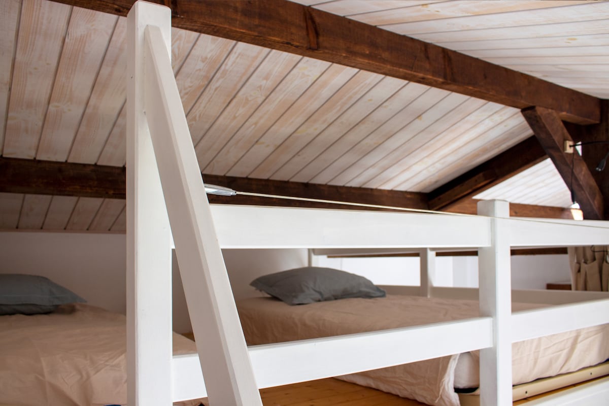 A cozy mezzanine area features two independent sleeping spaces, each with light bedding and soft pillows. The wooden ceilings and beams create a warm atmosphere, while a sturdy ladder provides access. The overall design emphasizes a comfortable and practical sleeping arrangement.