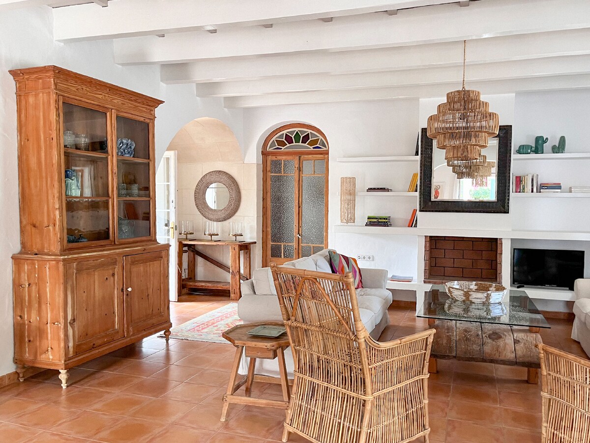 A spacious living room is characterized by natural light flowing through arched windows. A wooden display cabinet stands elegantly against one wall, while a cozy seating area includes wicker chairs and a glass coffee table. Decorative elements add warmth to the inviting space.