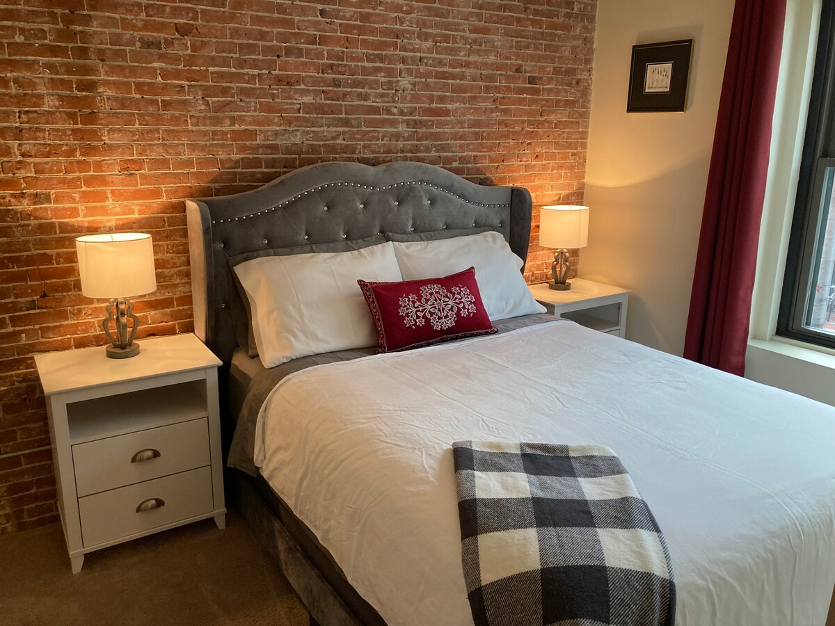 The bedroom features a queen size bed with a tufted gray headboard and white cotton linens. Two bedside tables with lamps flank the bed, and a decorative red pillow adds a pop of color. Exposed brick walls contribute to the room's character, complemented by natural light from a nearby window.
