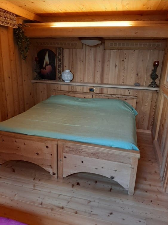 A cozy sleeping area features a wooden bed with a soft, light blue coverlet. The walls are adorned with warm wooden paneling, and a decorative candle painting adds a calming touch. Soft illumination highlights the serene atmosphere, ensuring a restful environment.