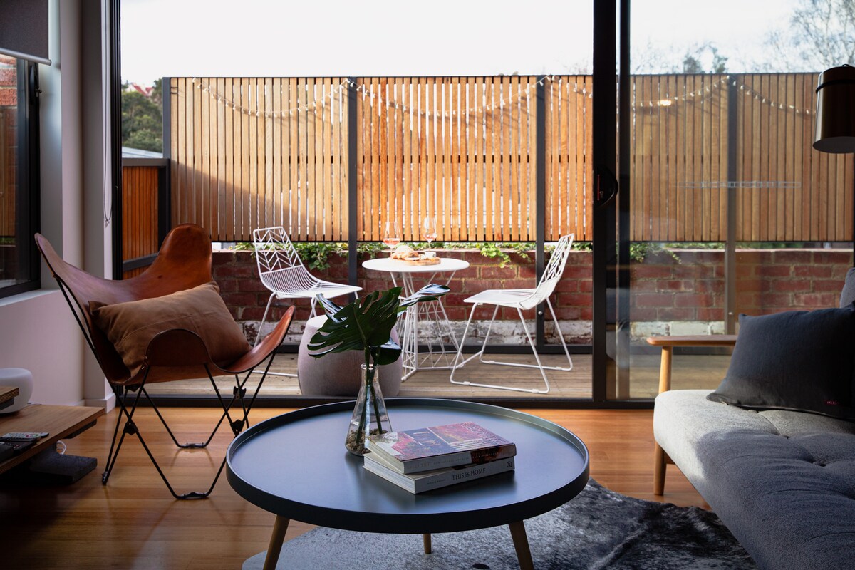 The image captures a bright living space featuring a round coffee table surrounded by modern outdoor furniture on a private deck. Large glass doors open to a fenced area with wooden materials, enhancing the connection with the outdoor environment. Natural light floods the room.