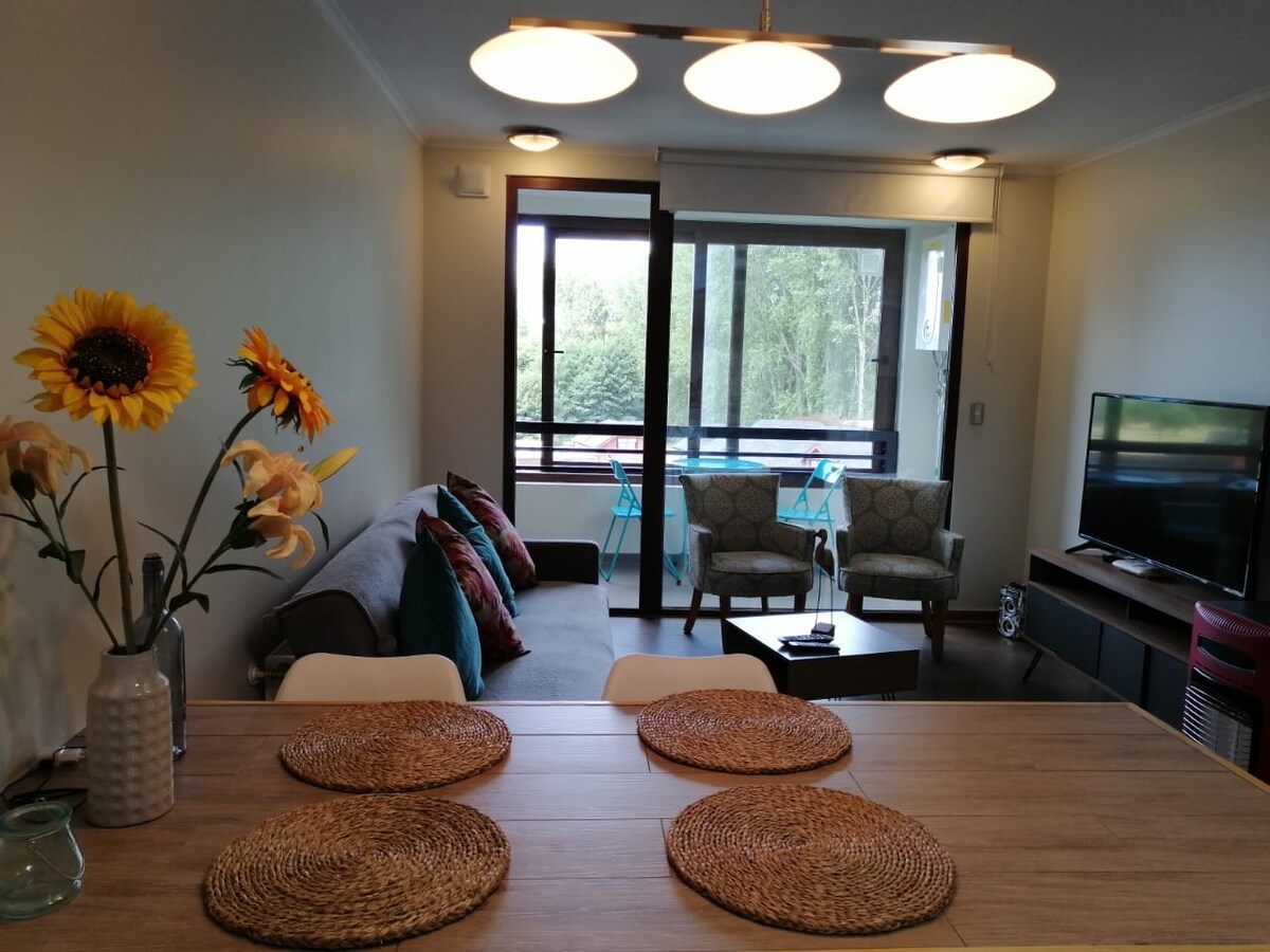 A modern living space features large windows providing natural light and views of greenery. Comfortable seating is arranged around a coffee table, with a television nearby. A dining table with woven placemats and a vase of flowers adds a welcoming touch to the room.