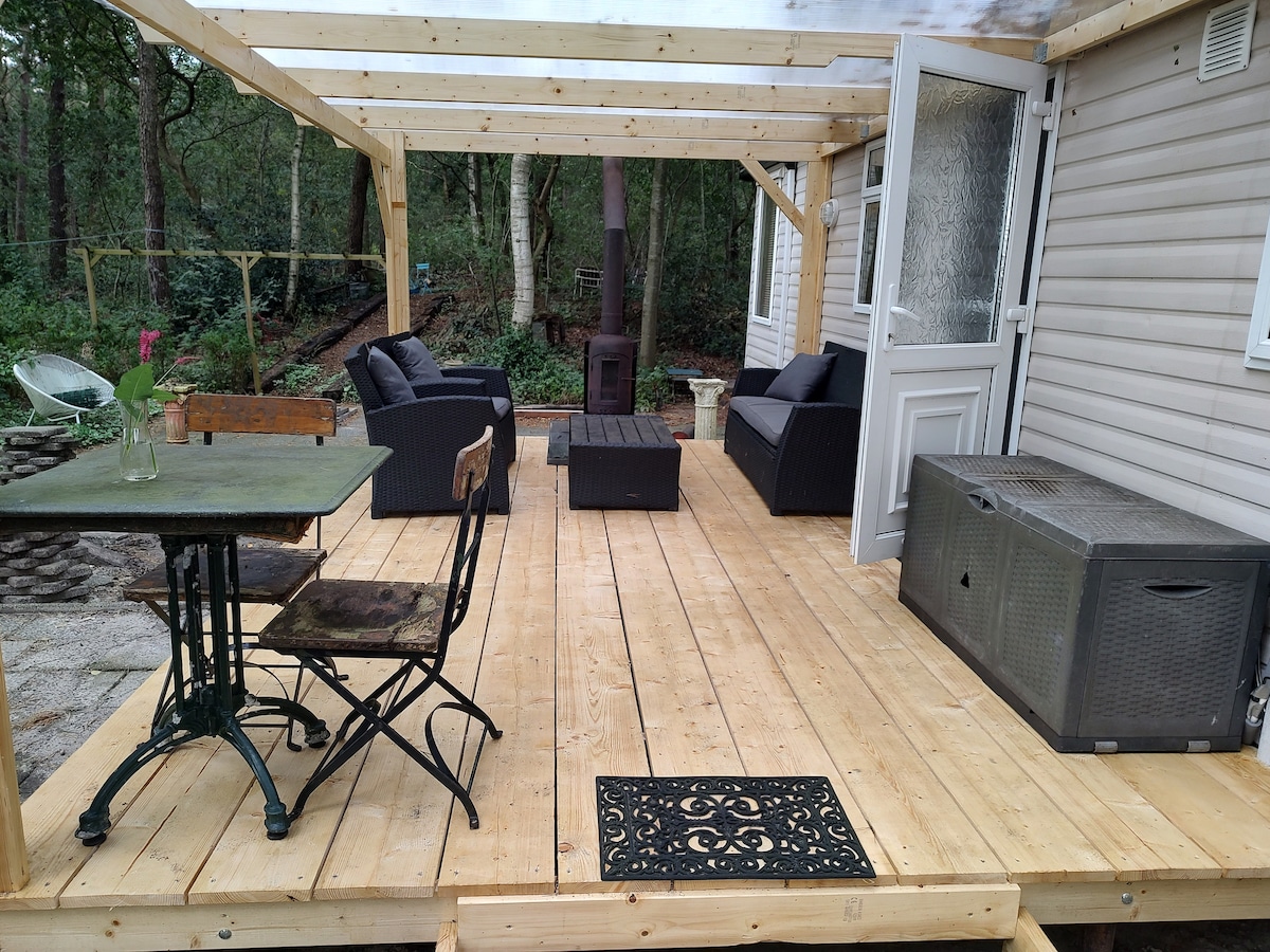 A spacious wooden terrace is shown, featuring a seating area with dark wicker chairs and a small table. A decorative rug is placed at the entrance, and a wooden stove is visible in the background. The surrounding area includes a natural setting with greenery.