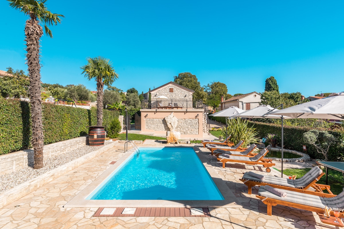 The outdoor area features a clear blue pool surrounded by lounge chairs, palm trees, and umbrellas. Lush greenery borders the space, creating a tranquil environment. A stone villa is visible in the background, enhancing the serene atmosphere of the property.