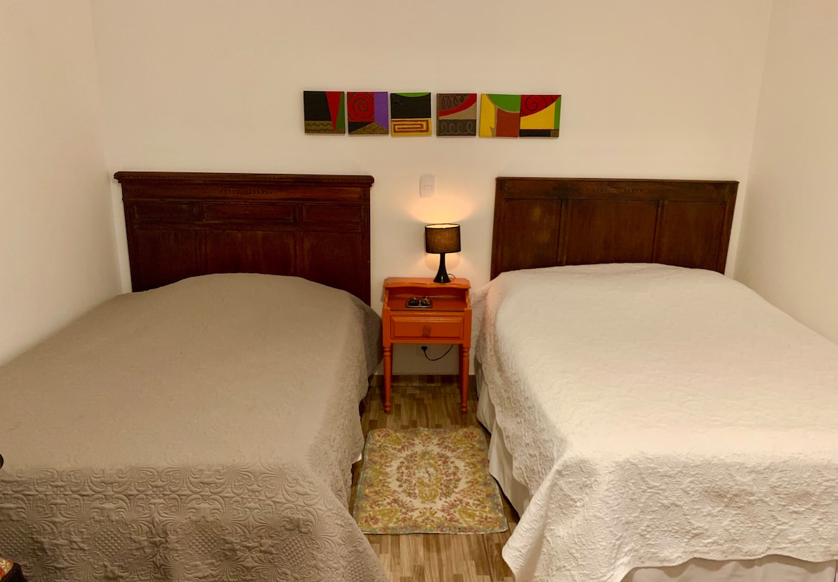 A well-lit sleeping area features two spacious beds, each topped with light-coloured bedding. An orange nightstand is positioned between them, holding a small lamp. A decorative area rug adds warmth to the wood floor, complemented by a colorful wall art piece above the beds.