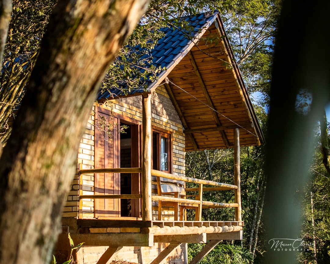 A charming wooden cabin is perched on stilts, featuring a sloped roof and a balcony that offers scenic views. Large windows are visible, allowing natural light to fill the interior. Surrounding trees add to the serene setting of the structure.