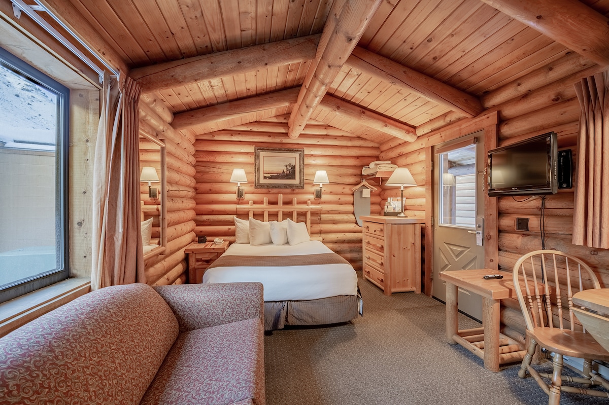 The interior of a log cabin is featured, showcasing wood-paneled walls and a ceiling with exposed beams. A queen bed is positioned against the wall, paired with nightstands. A seating area is provided with a sofa and a small dining table. Windows allow natural light to enter.