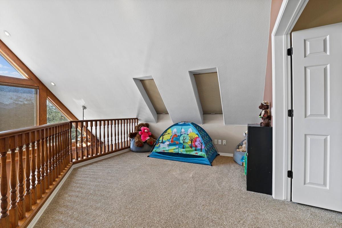 The loft space features soft carpet flooring and a sloped ceiling, allowing natural light to enter through skylights. A colorful play tent is positioned in the corner, surrounded by plush toys. A wooden railing provides an overlook to the main level.