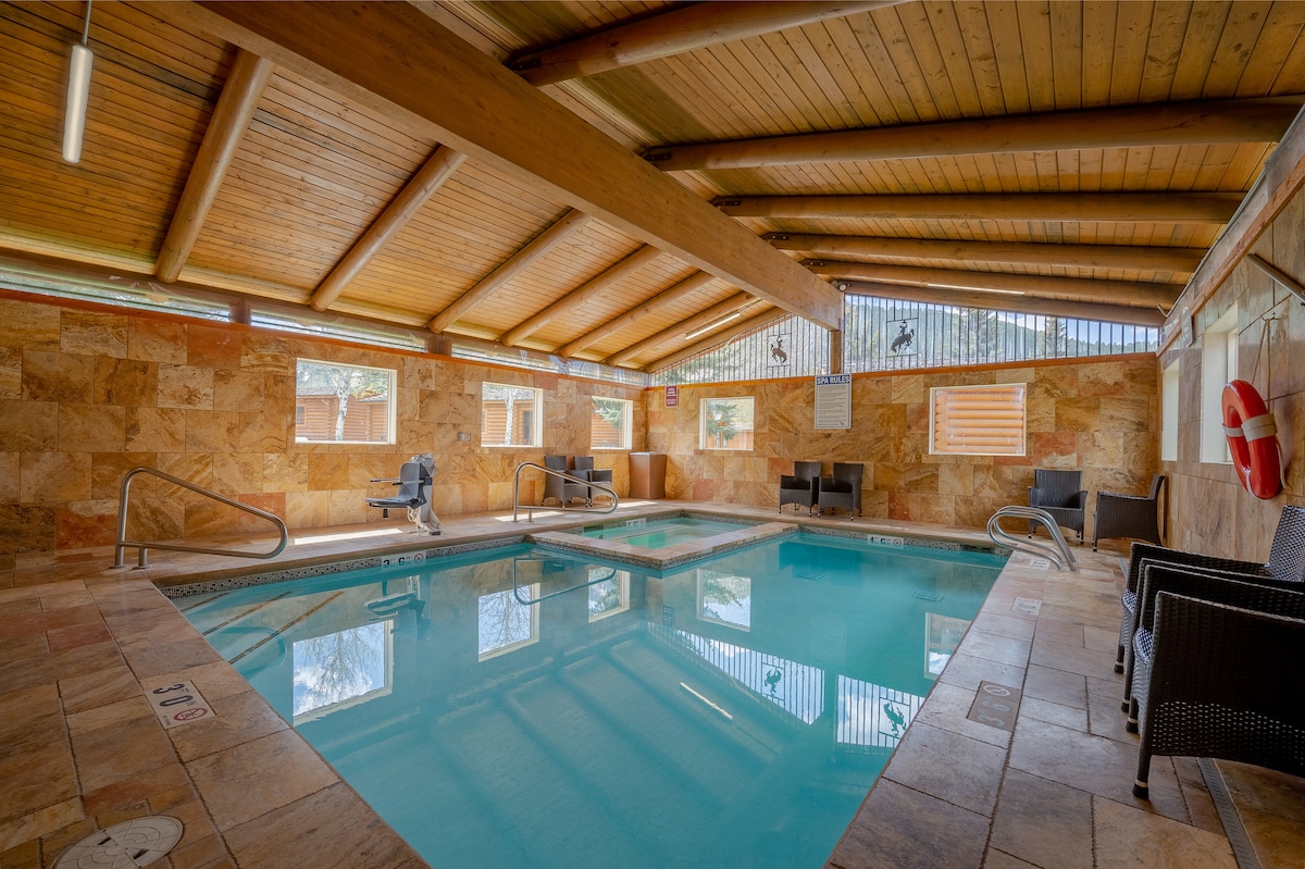 An indoor pool area features a rectangular pool surrounded by warm stone walls and wooden beams. Lounge chairs are positioned near the water, and large windows allow natural light to illuminate the space.