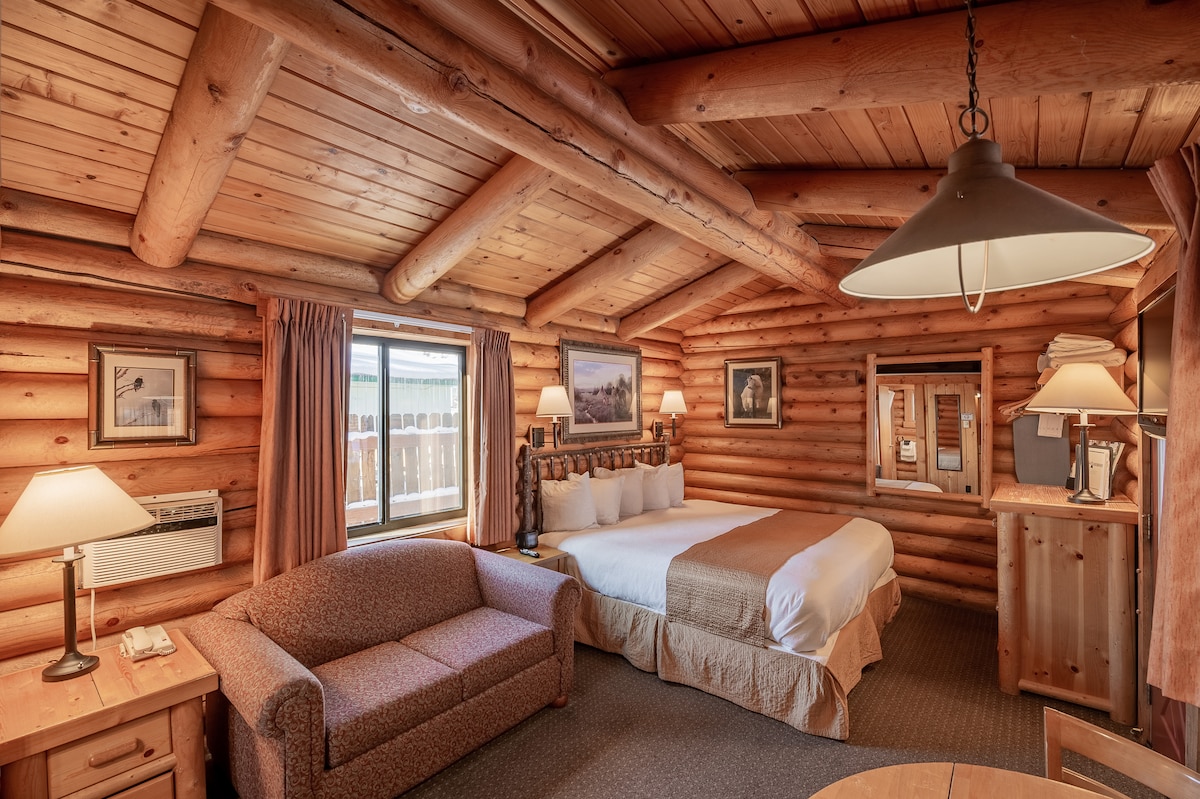 A log cabin interior features warm wood paneling, with a spacious bed and a sofa situated nearby. Large windows allow natural light to fill the room. A small dining area is visible, accompanied by lamps that provide soft lighting.