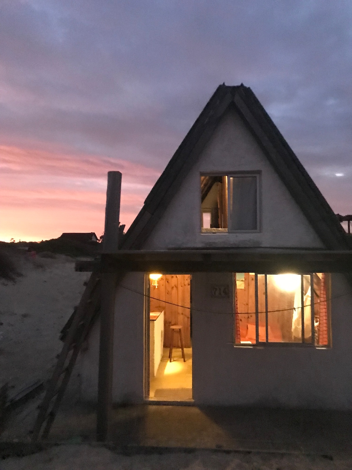 The triangular structure features a rustic exterior with wooden accents and a warm glow emanating from the windows. A sandy pathway leads to the entrance, complementing the colorful sunset sky in the background.