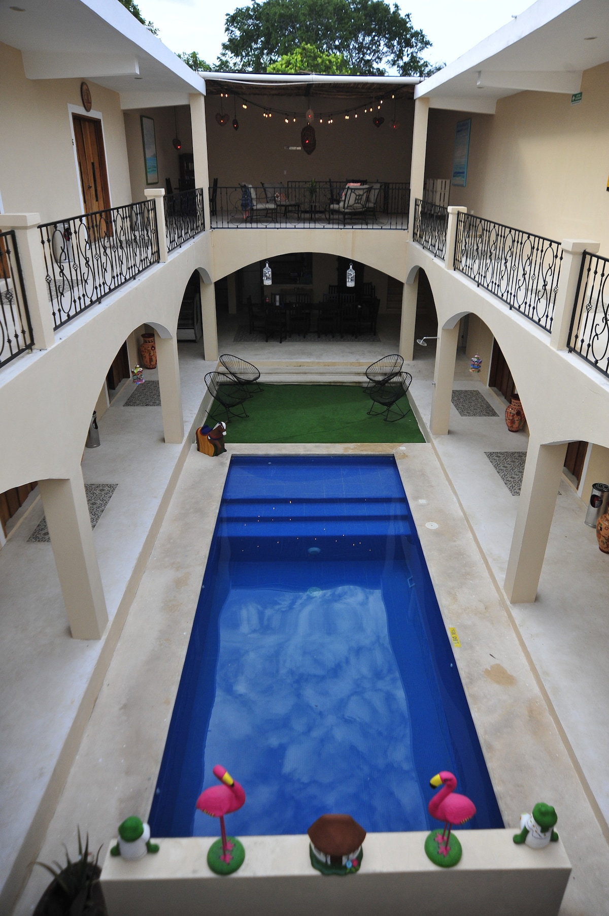 A central pool surrounded by a patio area features a seamless blue surface reflecting the sky. Arched balconies overlook the pool, with decorative elements enhancing the surroundings. Lounge chairs are positioned on a grassy area, providing a space for relaxation.