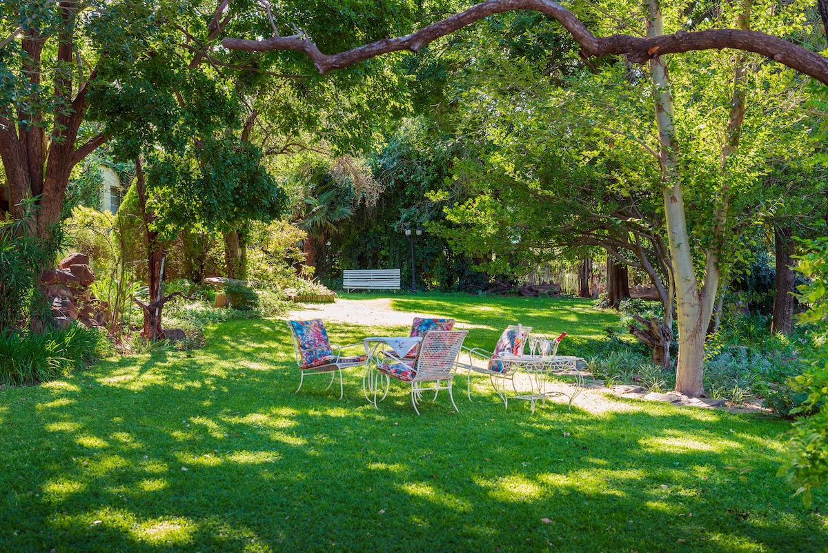 A lush garden is presented with vibrant greenery, featuring several chairs arranged around a round table. Sunlight filters through the trees, creating playful patterns on the grass. A white bench is visible in the background, adding to the inviting outdoor atmosphere.