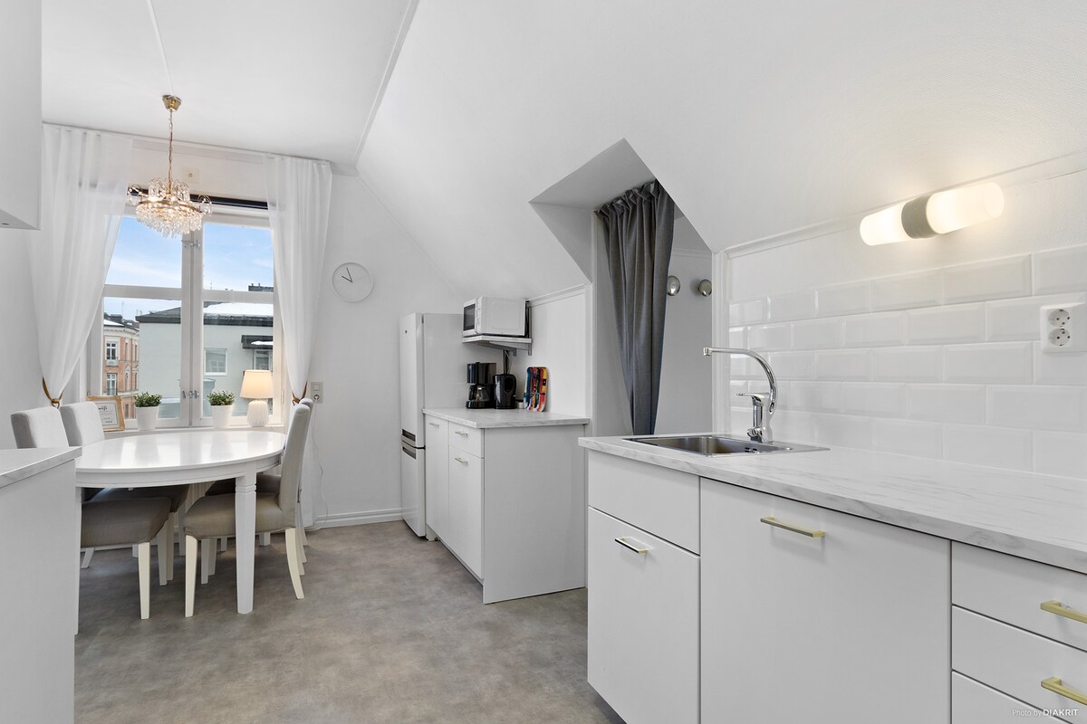 A bright kitchen with modern appliances and a dining area. A round table surrounded by white chairs is positioned near a window, allowing natural light to fill the space. The sleek countertops and cabinetry contribute to a clean and functional atmosphere.