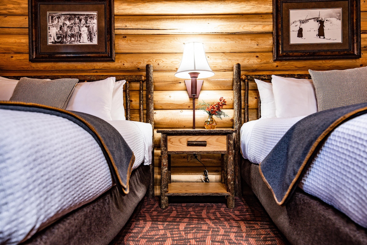 The cozy sleeping area features two queen beds, framed by natural wood logs. A bedside table made of wood is positioned between the beds, with a lamp providing soft illumination. Historic photographs decorate the walls, enhancing the rustic ambiance.