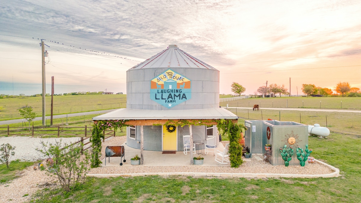 The exterior of The Silo House is showcased at sunset, with a gray metal structure adorned with vibrant signage. Rocking chairs are arranged on the front porch, and a small lawn area with decorative plants is visible. The surrounding landscape is open, with grazing animals in the distance.