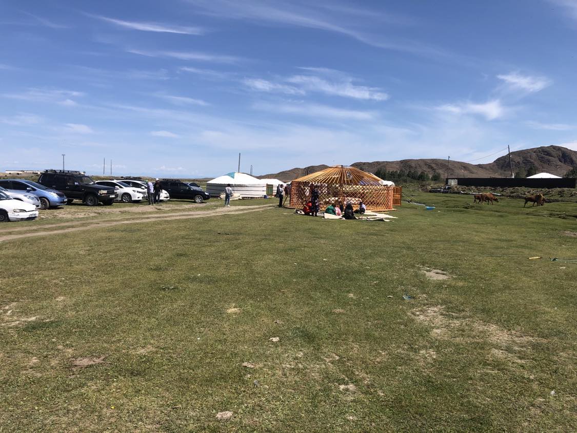 A spacious outdoor area is shown, featuring a traditional yurt surrounded by green grass. Parked vehicles are visible in the background, while people are gathered on the ground nearby. Rolling hills are seen in the distance under a clear blue sky.