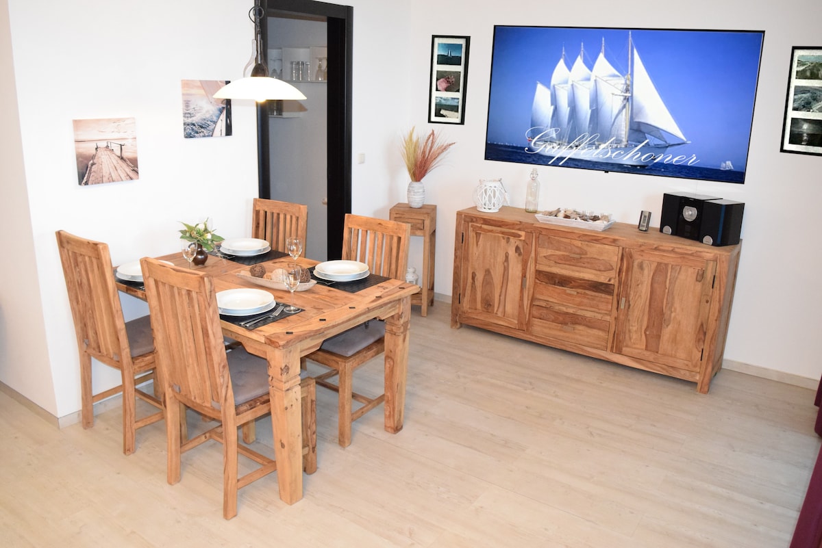 A dining area features a wooden table with four chairs, set for a meal with plates and glassware. A large wall-mounted television displays an image of sailboats, while a sideboard provides additional storage and decor, enhancing the room's modern aesthetic.