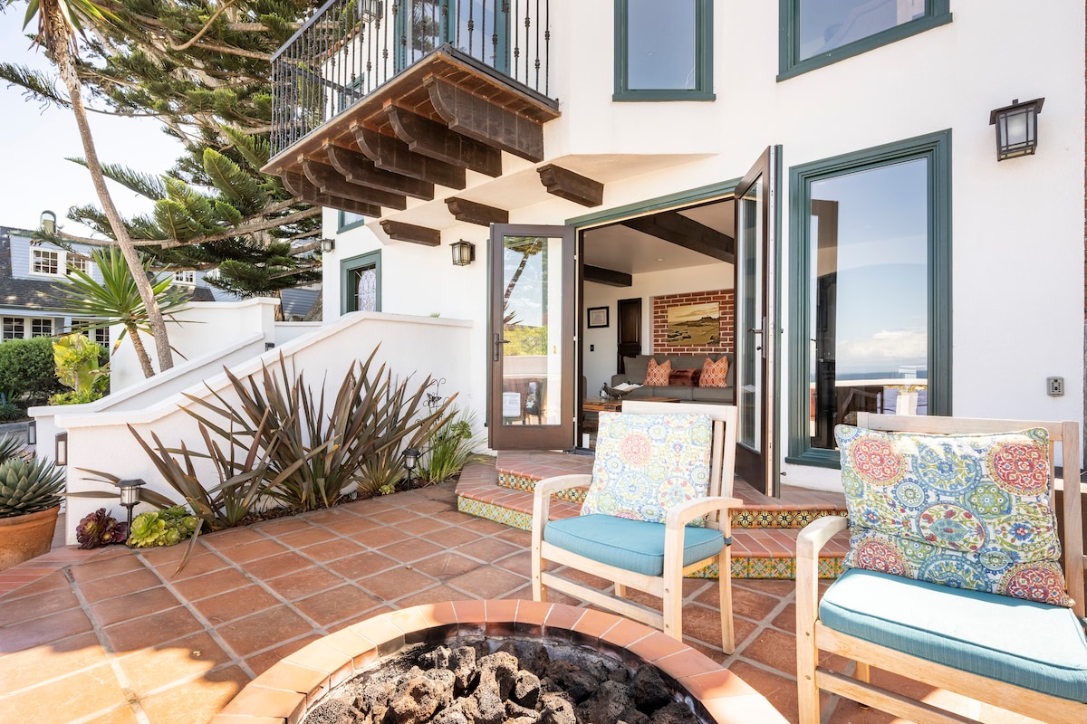 An outdoor patio features a fire pit surrounded by comfortable seating with colorful cushions. Terracotta tiles create a warm atmosphere, complemented by surrounding greenery and ocean views. Large glass doors open into the interior living space, inviting natural light inside.
