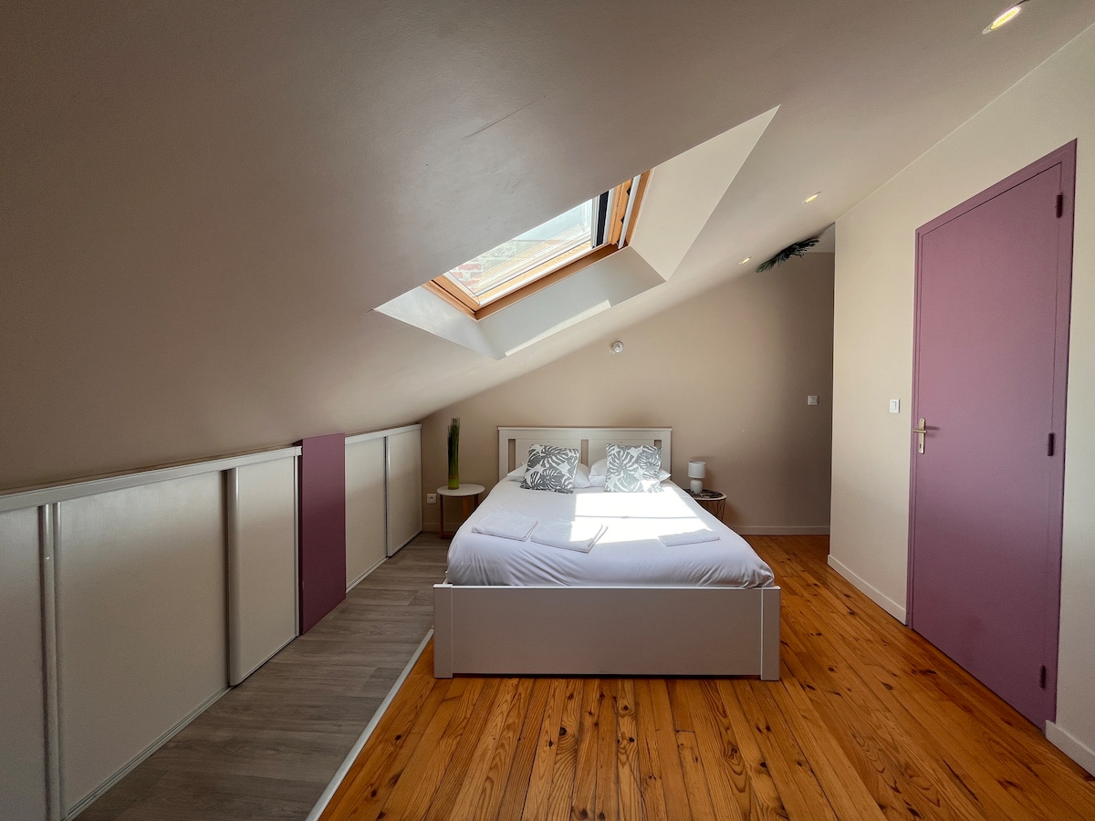A spacious bedroom features a comfortable double bed with decorative pillows. Natural light enters through a skylight, highlighting the warm wooden flooring. Storage cabinets line the walls, complemented by a subtle color palette. A side table with a lamp adds functional detail to the serene space.