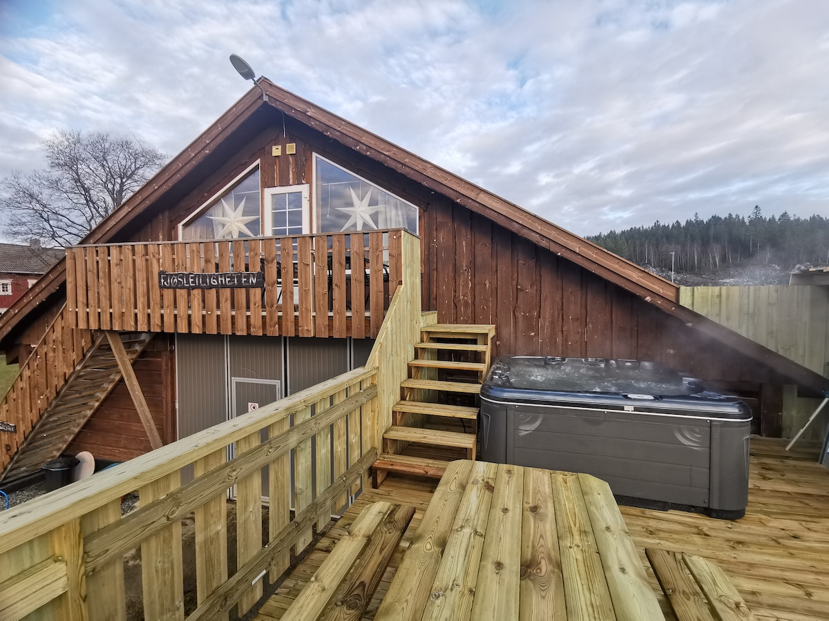 A wooden terrace features a jacuzzi and leads to the entrance of the upper-level apartment. The rustic facade is characterized by large windows and a steeply pitched roof, surrounded by trees in the background.