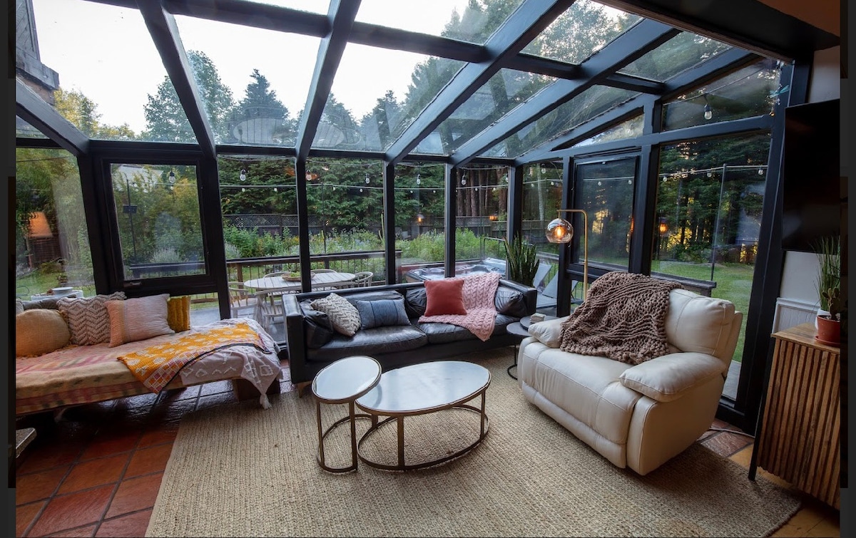 A sunlit sunroom features a mix of modern and cozy furnishings, including a white leather sofa and a neutral-toned rug. Large glass panels frame views of the surrounding greenery, while comfortable throws are arranged on the seating. A round coffee table sits centrally, inviting relaxation.