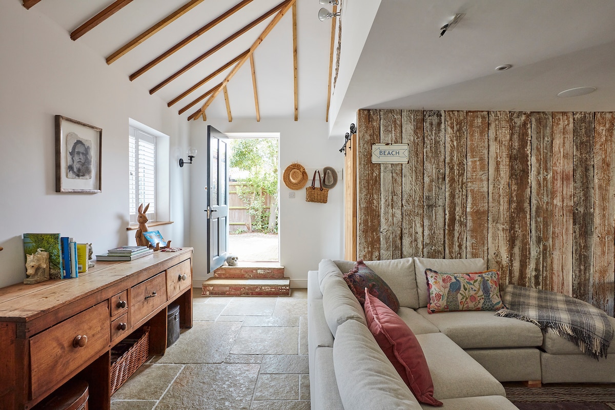 A living area is presented with natural stone flooring and exposed wooden beams on the ceiling. A light-colored sofa is positioned against a rustic wooden wall. Nearby, a door opens to a garden area, complemented by decorative items on a wooden sideboard.