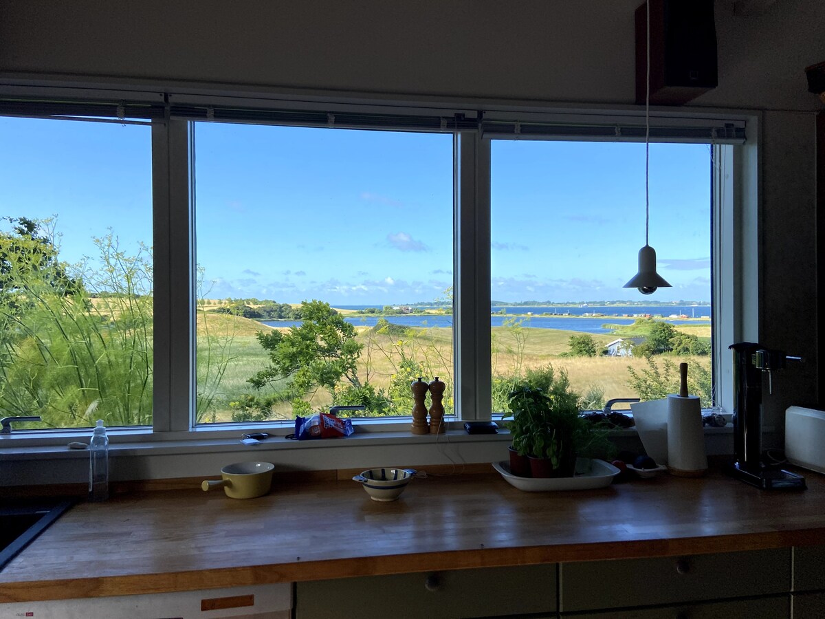 A well-appointed kitchen features a spacious wooden countertop with various kitchen utensils. Large windows provide a clear view of the lush landscape and water, inviting natural light into the space.