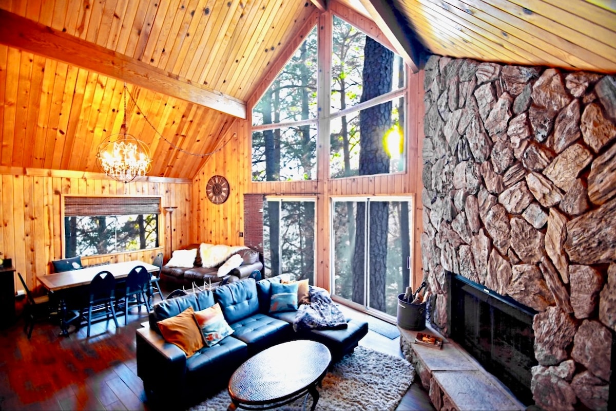 An inviting living space showcases a combination of wooden walls and vaulted ceilings. Large windows provide views of surrounding trees. A stone fireplace stands prominently, while a plush black sofa is adorned with colorful pillows. A cozy rug lies beneath a round coffee table, enhancing comfort.