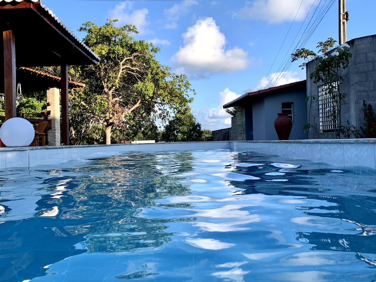 A clear blue pool reflects the sky with fluffy clouds. Surrounding greenery adds a natural touch. A shaded pavilion with wooden seating is visible in the background, along with a neighboring house that features a terracotta pot beside it.
