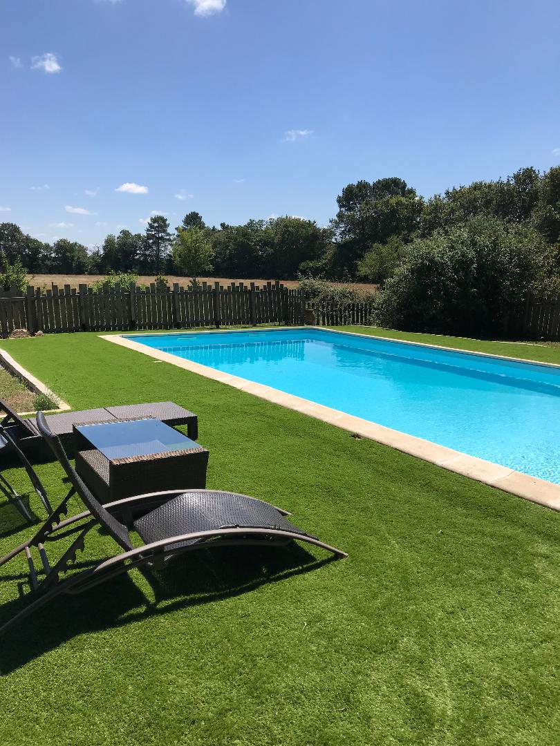 A private salt-water pool is surrounded by well-maintained green grass. Lounge chairs are positioned nearby, providing a comfortable space to relax. Tall trees and a wooden fence create a secluded natural setting, with a clear blue sky overhead.