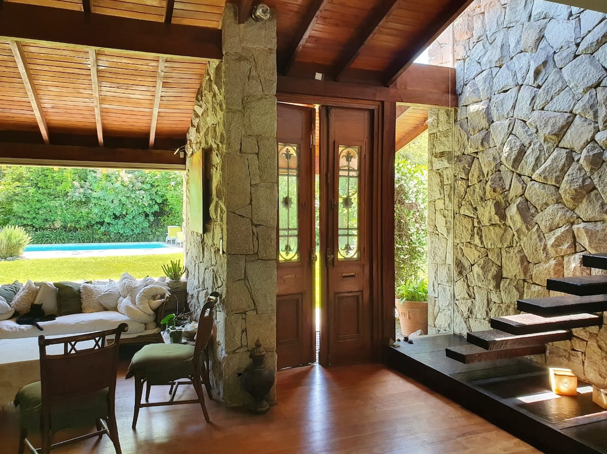 The entryway features double wooden doors framed by stone walls and natural light streaming in. A comfortable seating area is visible beside a large window, and a staircase leads to the upper level, complemented by a wooden ceiling and a warm wooden floor.