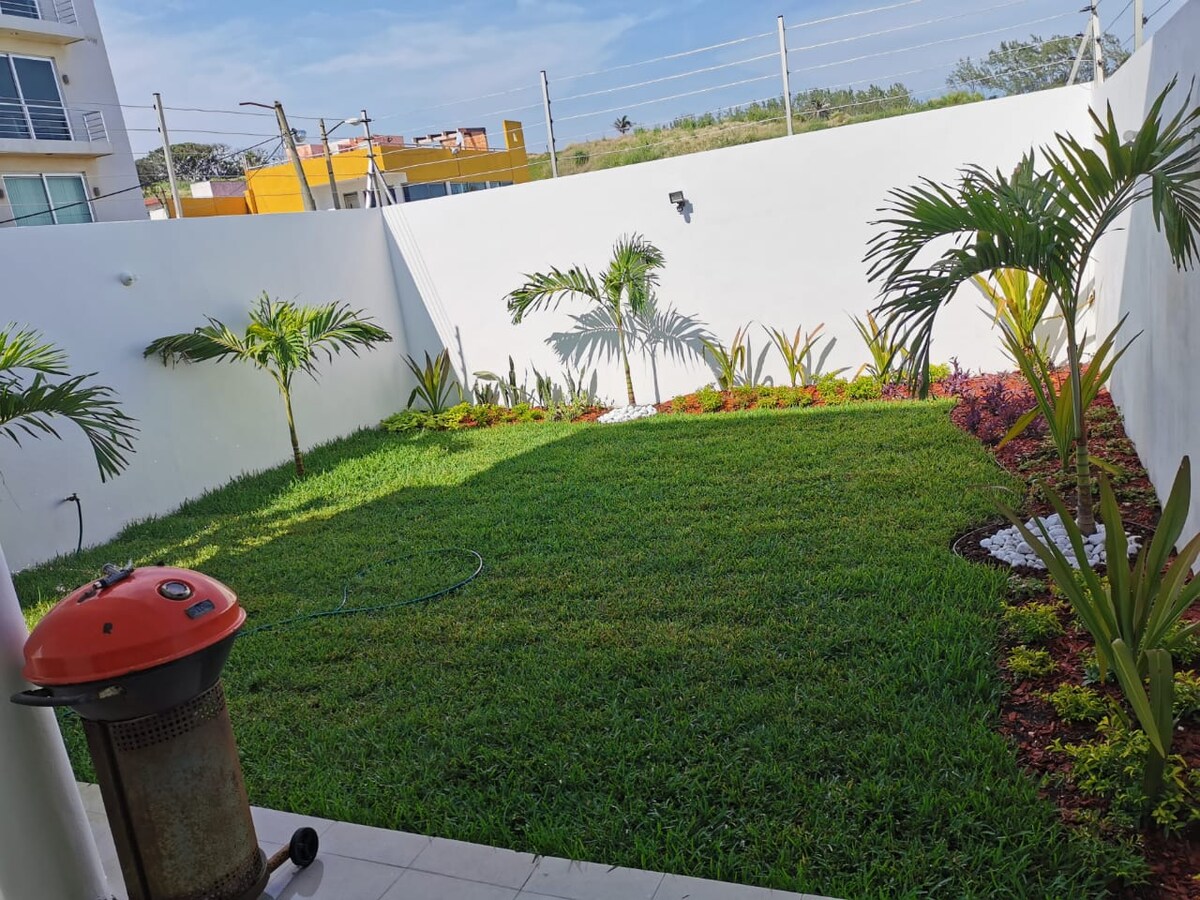 A well-maintained garden area features lush green grass and neatly arranged flower beds with colorful plants. Small palm trees are positioned throughout the space, providing a tropical feel. A barbecue grill is placed on one side, adding functionality to the outdoor setting.