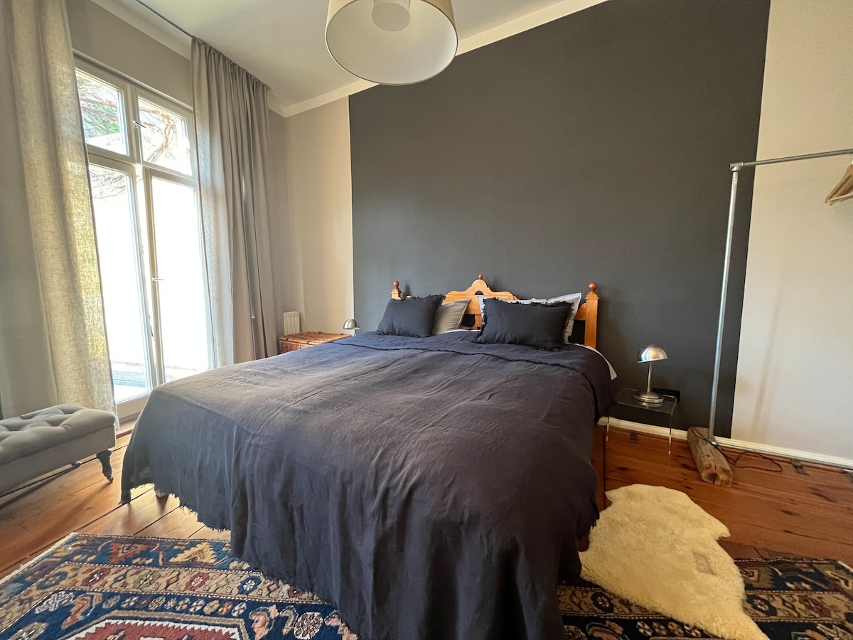 A spacious sleeping area features a large, dark bed adorned with matching pillows. A stylish bedside lamp and a wooden dresser complement the room. Natural light enters through large windows, framed by light curtains, while an area rug adds a touch of warmth to the wooden floor.