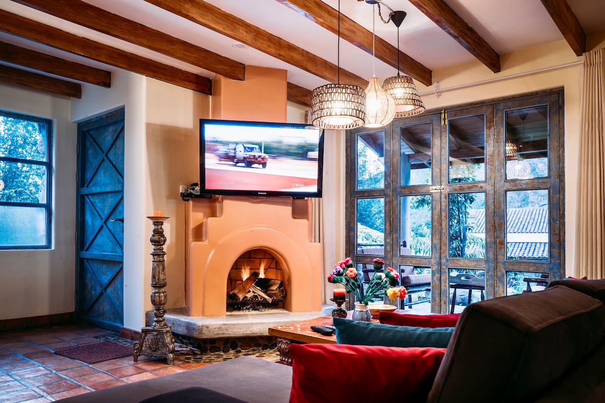 A comfortable living area features a fireplace surrounded by stone, with a large television mounted above. Wooden beams cross the ceiling, and large windows offer views of the outdoors. A couch with colorful cushions complements a coffee table adorned with fresh flowers.