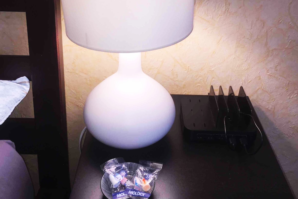 A bedside table is adorned with a softly glowing lamp featuring a round base. Nearby, a small plate holds complimentary candies. A router and a set of charging stations are positioned on the table, providing convenience for electronic devices.