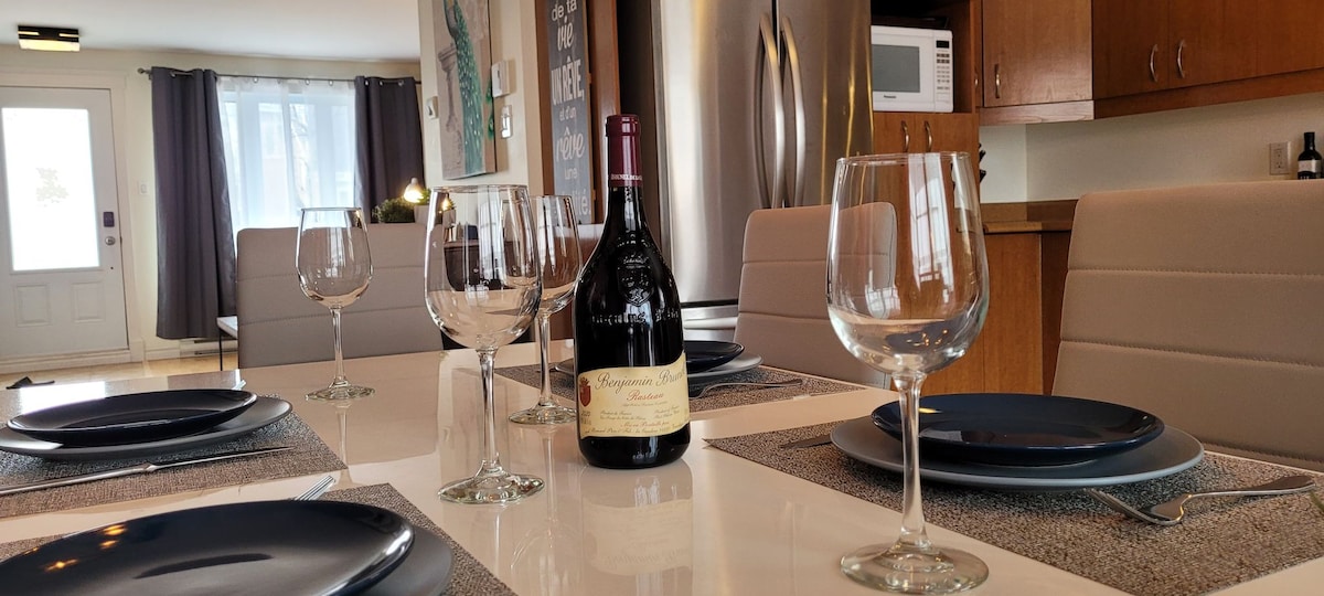 A modern dining area is presented, featuring a white table set with dark plates and crystal wine glasses. A bottle of wine is centrally placed. Behind, sleek appliances and minimalist cabinetry enhance the kitchen's contemporary design, while natural light brightens the space through the front door.