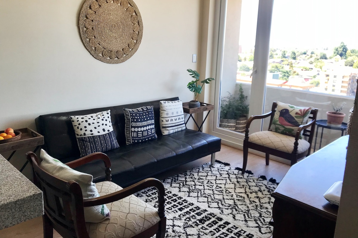 A spacious living area features a modern black sofa adorned with decorative cushions. Two wooden side tables frame the seating, while a potted plant adds a touch of greenery. Light filters through large glass doors that lead to a balcony with urban views.