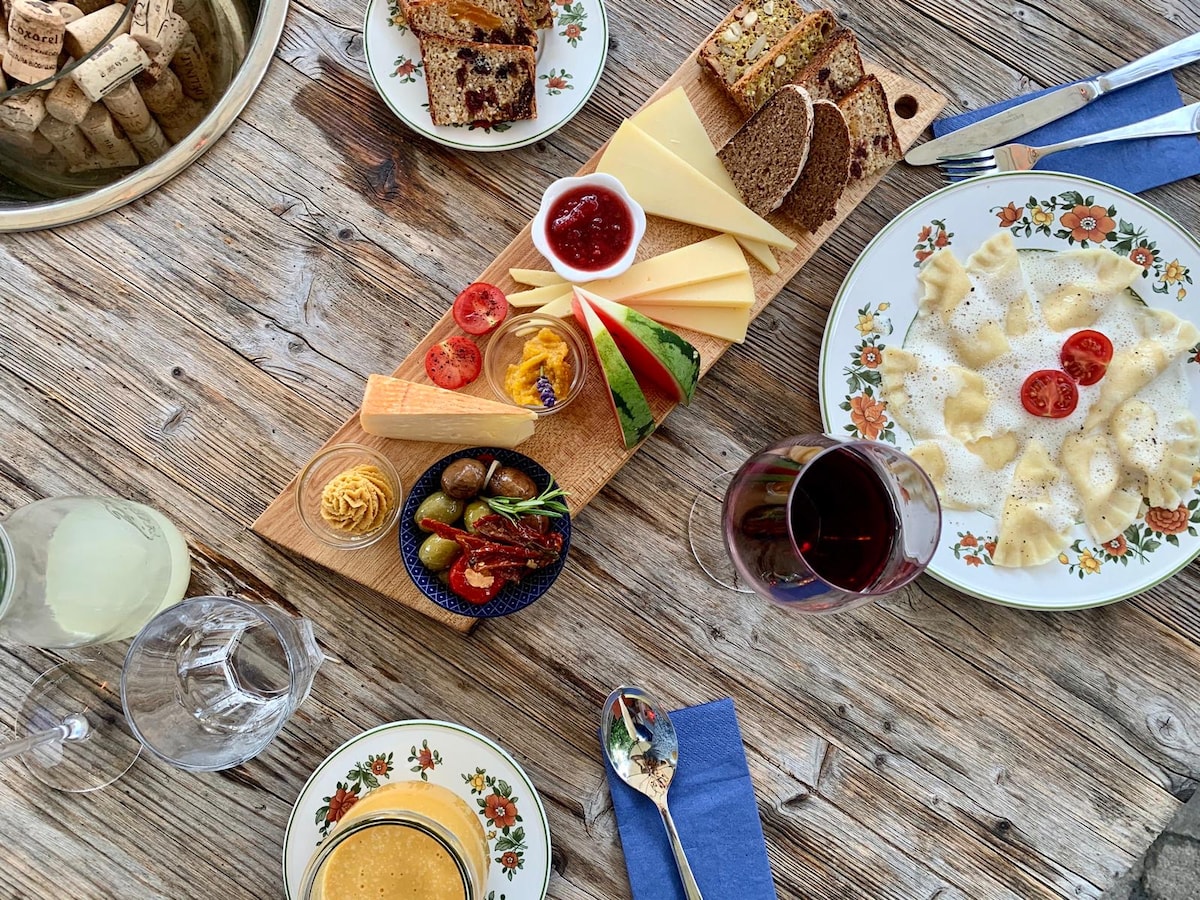 A wooden serving board displays a selection of cheeses, breads, and jam, accompanied by a small dish of olives. Plates with pasta drizzled in sauce are seen alongside glasses containing beverages, all set on a rustic wooden table with floral-patterned dishware.
